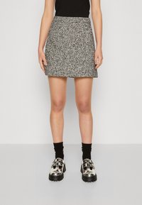 Žena v černobílé texturované mini sukni, černých ponožkách, lesklých stříbrných botách na přezku a černém topu bez rukávů na bílém pozadí.