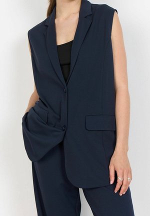 Blazer bleu marine sans manches porté sur un top noir, associé à un pantalon bleu marine assorti ; main dans la poche du blazer, bagues visibles aux doigts.