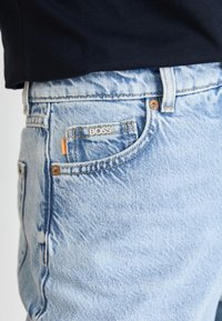 Jasnoniebieskie dżinsy z logo BOSS na przedniej kieszeni. Oferują klasyczne szwy i mały pomarańczowy akcent przy kieszeni.