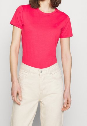 Kobieta ubrana w jasnoróżową, krótkim rękawem, t-shirt z okrągłym dekoltem oraz wysokim stanem kremowe spodnie na jednolitym tle.