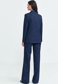 Marynarski garnitur w paski wykonany z gładkiego materiału. Posiada dopasowaną marynarkę oraz szerokie spodnie, z nałożonymi guzikami na rękawach marynarki.