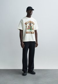 Ανετη κρεμ μπλούζα με σχέδιο αρκούδας και κείμενο "Billionaire Boys Club", συνδυασμένη με μαύρο παντελόνι και μαύρα αθλητικά παπούτσια.