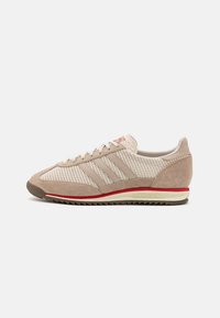 Zapatilla deportiva beige con parte superior de malla texturizada, acentos de ante y tres rayas en contraste. Cuenta con una suela de goma con detalles rojos.