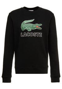 Černá mikina s logem zeleného krokodýla a textem "LACOSTE" v tučných bílých písmenech. Vyrobena z měkké, strukturované látky s kulatým výstřihem.