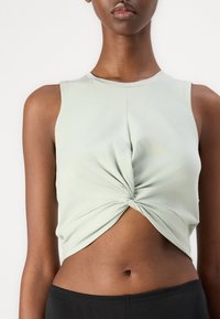 Svijetlo zelena, bez rukava, kratka majica s preokrenutim prednjim dizajnom; glatka tekstura i prilagođen oblik. Uparen s crnim hlačama.