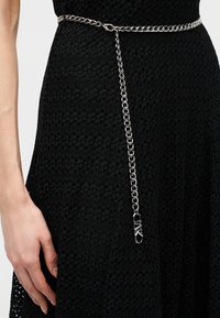 Ceinture chaîne argentée avec une boucle en forme de boucle suspendue sur une robe noire texturée à la taille, avec une main reposant à côté de la jupe.