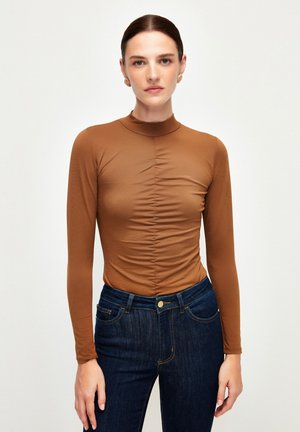 Maglia marrone a maniche lunghe con colletto alto e dettagli arricciati sul davanti, abbinata a jeans di denim blu scuro con un classico design a cinque tasche.