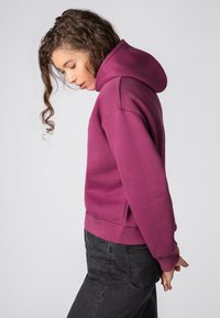 Diepe paarse pullover hoodie van stof met een voorzak en geribbelde zoom. Beschikt over een grote, aangesloten capuchon en een ontspannen pasvorm. Gekleed met zwarte broek.