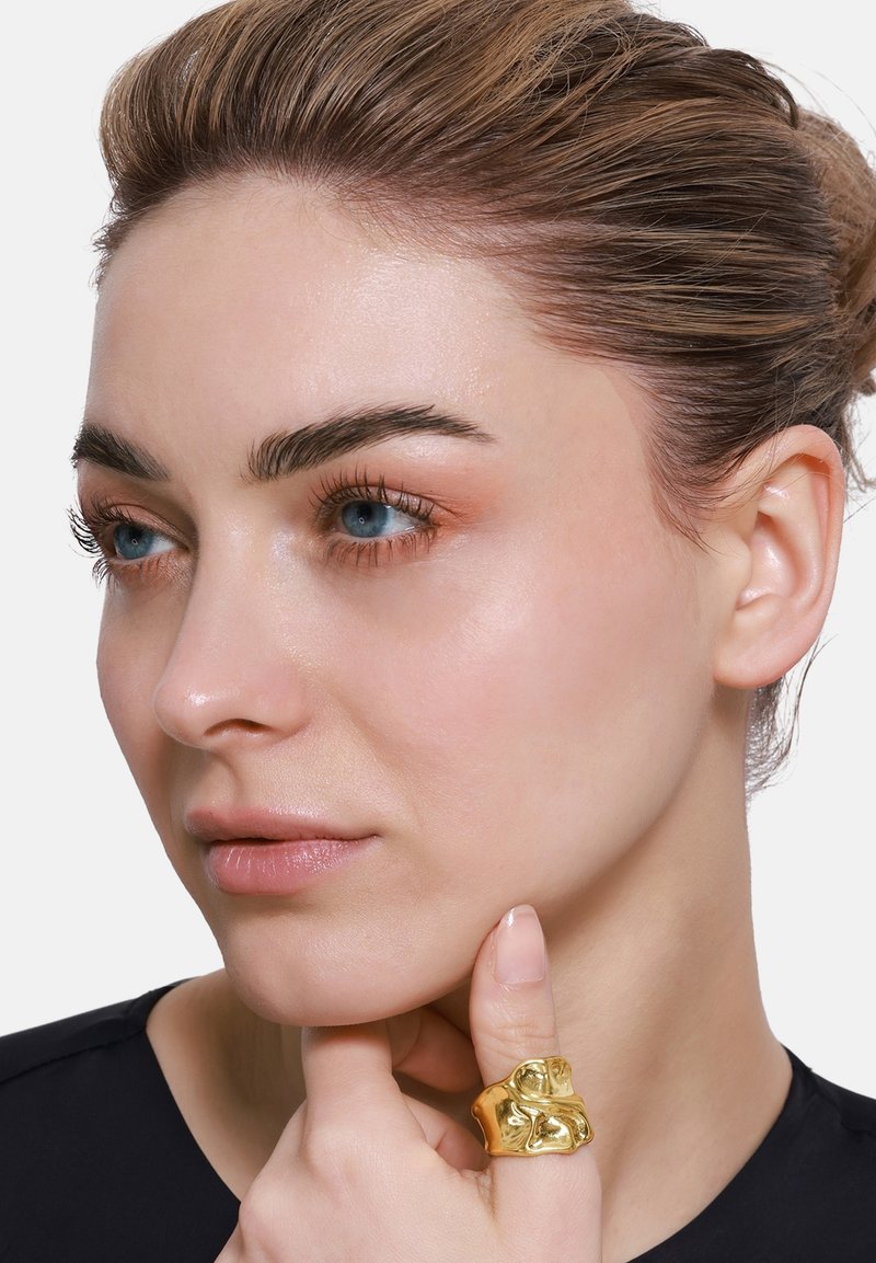 Gouden textuur ring om een vinger, met gladde lijnen en een sculpturaal ontwerp. Het model heeft neutrale make-up, blauwe ogen en een zwarte top.