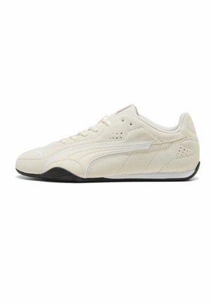 Zapatilla deportiva de ante beige con detalles blancos, suela negra, cierre de cordones y diseño bajo, mostrada de perfil sobre fondo blanco.