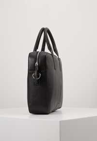 Mallette en cuir noir avec double poignée et fermeture éclair supérieure, debout verticalement sur une surface blanche contre un fond clair uni.