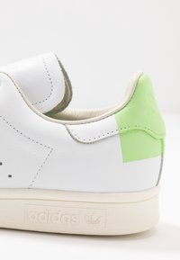 Vita Adidas-sneakers med ljusgrön hälpatch, beige sula och synliga sömmar på sidan och kragen.