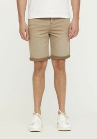 Beige Denim-Shorts mit umgeschlagenen Säumen, frontalen Taschen und sichtbaren Nähten. Getragen mit weißen Sneakern vor einem einfarbigen Hintergrund.
