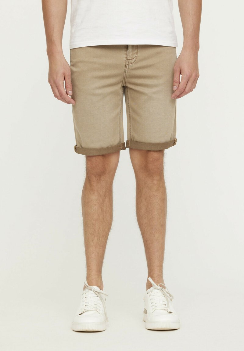 Beige Denim-Shorts mit umgeschlagenen Säumen, frontalen Taschen und sichtbaren Nähten. Getragen mit weißen Sneakern vor einem einfarbigen Hintergrund.