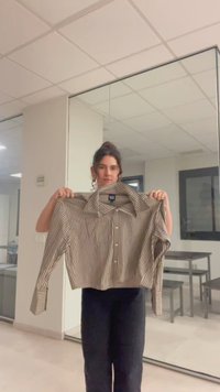 Femme tenant une chemise courte à manches longues, rayée beige et marron, boutonnée, dans une pièce lumineuse avec un grand miroir et des tables.