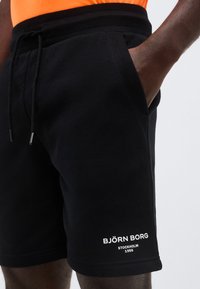 Άνδρας που φοράει μαύρα σορτς Björn Borg με πλαϊνές τσέπες, με το ένα χέρι μέσα στην τσέπη, συνδυασμένα με πορτοκαλί μπλουζάκι.