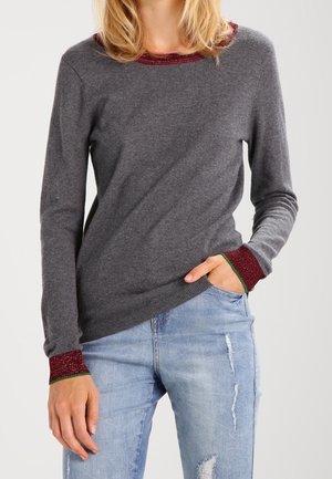 Femme portant un pull gris à manches longues avec des bordures rouges et vertes au col et aux manches, associé à un jean bleu clair déchiré.
