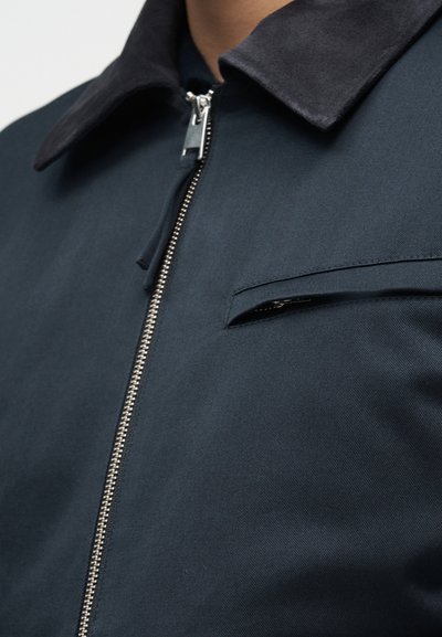 Σκούρο μπλε σακάκι με ασημένιο φερμουάρ στο μπροστινό μέρος, μαύρο γιακά και τσέπη στο στήθος με φερμουάρ στην αριστερή πλευρά.