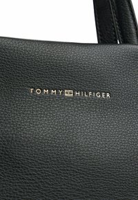 Sac à main en cuir noir avec une finition texturée. Le logo de la marque "TOMMY HILFIGER" est embossé en or sur le devant.