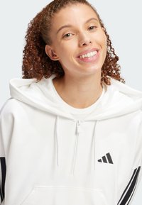 Weißer Hoodie mit Reißverschluss am Kragen, ausgestattet mit einer Kängurutasche und schwarzem adidas-Logo. Er hat schwarze Streifen an den Ärmeln.