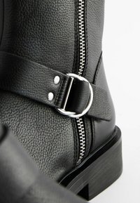 Gros plan d'une botte en cuir noir texturé avec fermeture éclair argentée, bride et boucle métallique ronde sur semelle à talon empilé.