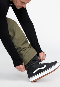 Chemise noire à manches longues, pantalon vert olive et baskets montantes noires avec des accents blancs. Une personne ajuste les poignets de son pantalon au-dessus de ses chaussures.