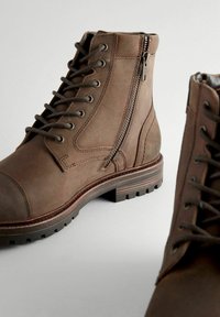 Botas de tobillo de cuero marrón con cordones y cremallera lateral, punta redondeada, superficie texturizada y suela de goma con surcos para tracción.