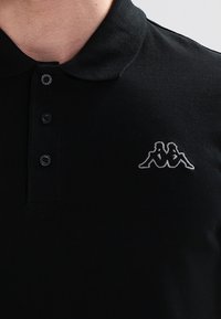 Svart pikétröja i bomullstyg; har en treknappsknäppning och en broderad logotyp på vänster bröst.