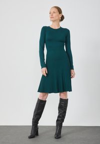 Rochie tricotată teal cu mâneci lungi, textură côtelată și fustă plisată evazată, asortată cu botine negre până la genunchi. Decoltare rotundă simplă.