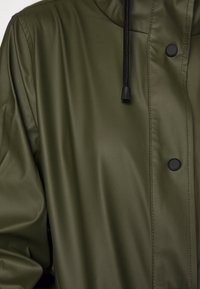 Giacca impermeabile verde oliva in materiale liscio e lucido; dotata di cappuccio, coulisse nera e bottoni a pressione neri.