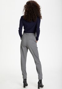 Femme aux cheveux bouclés portant un haut bleu marine à manches longues, un pantalon gris ajusté et des bottines à talons noires, debout les mains dans les poches.