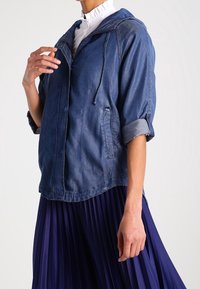 Giacca di jeans blu scuro con zip frontale e cappuccio regolabile. Presenta maniche arrotolate e due tasche laterali, abbinata a una gonna pieghettata viola.