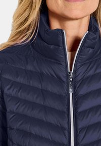 Marine blauwe gewatteerde jas gemaakt van synthetisch materiaal; heeft een hoge kraag, een rits aan de voorkant en diagonale stiksels voor isolatie.