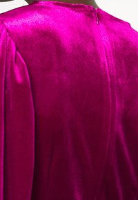 Vêtement en velours fuchsia avec une texture lisse. Doté de manches douces et plissées et d'une fermeture éclair discrète dans le dos.