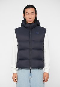 Tums vāciņš tumši zils, ar kapuci, priekšējo rāvējslēdzēju un zīmola uzlauku. Audums izskatās gluds ar quiltētu, pildītu dizainu.