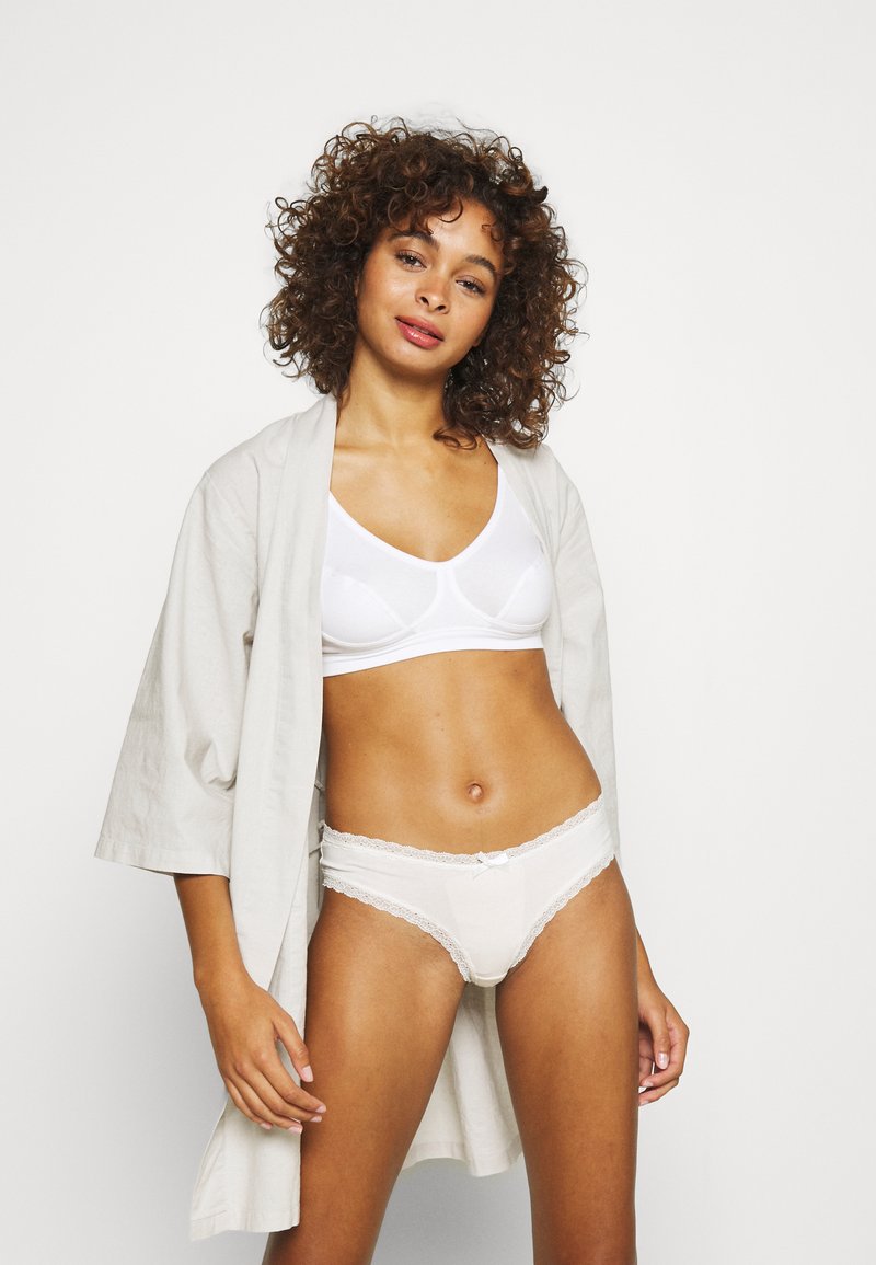 Bralette in cotone bianco e mutandine con bordi in pizzo, sovrapposte a un kimono beige chiaro. Texture morbide, tonalità tenui, vestibilità e design informale.