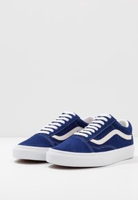 Baskets en suède bleu avec des lacets blancs, arborant un accent en forme de vague blanche sur le côté et une semelle en caoutchouc texturée. Design classique bas.