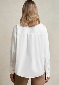 Cămașă albă cu mânecă lungă, hem rotunjit, detaliu cu pliu la spate și mansete cu nasturi, purtată cu pantaloni crem deschis. Textură de țesătură moale.