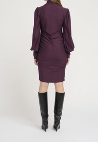 Fialové těsně přiléhavé šaty s naddimenzovanými rukávy a vysokým límcem, s nařasenými detaily na zádech, v kombinaci s černými kozačkami na podpatku sahajícími po kolena.