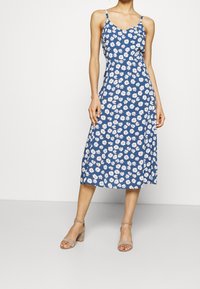 Robe fleurie bleue avec des marguerites blanches, dotée de fines bretelles, d'un corsage ajusté et d'une jupe mi-longue. Portée avec des chaussures à talons carrés beiges.