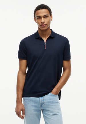 Homme portant un polo bleu marine à manches courtes avec col zippé et un jean bleu clair, debout devant un fond blanc uni.