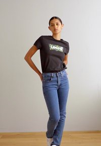 Melna T-krekli ar zaļo kamuflāžas Levi's logo priekšpusē, apģērbs apvienots ar gaiši ziliem džinsiem un baltiem sporta apaviem, stāvot uz neitrāla fona.