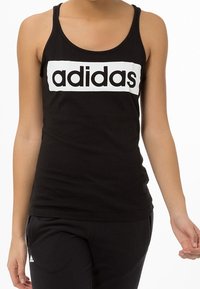 Czarny top na ramiączkach wykonany z bawełny, z okrągłym dekoltem, z białym prostokątnym paskiem z logo "adidas" w czerni.