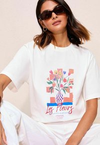 Weißes Oversized-T-Shirt mit einem bunten Grafik eines Blumenvasens, in lebhaften Rottönen, Rosa und Lila, präsentiert vor einem gemusterten Hintergrund.
