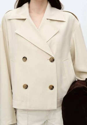 Flødefarvet dobbeltradet jakke med store revers og mørke knapper, båret over matchende bukser, person der holder en mørkebrun taske.