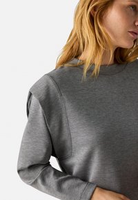 Sudadera gris con cuello redondo y mangas largas. Presenta una textura suave y costuras de hombro estructuradas. Diseño simple y minimalista.