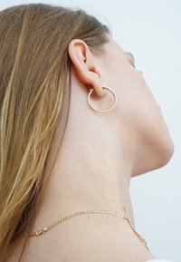 Goldener Creolenohrring, der am Ohr eines Models befestigt ist, mit einem glatten, runden Design. Das Model trägt eine filigrane goldene Halskette.