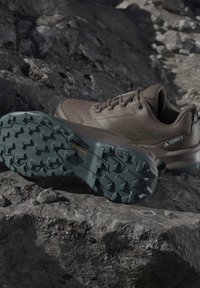 Brązowe buty trekkingowe Adidas Terrex z głębokim bieżnikiem Continental na podeszwach z gumy leżące na skalistym terenie.
