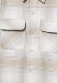 Plaid shirt in soft beige and white fabric, featuring two chest pockets, rounded collar, and light-colored snap buttons.