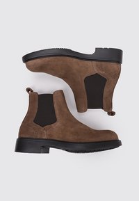 Botas de tobillo de ante marrón con paneles elásticos en los laterales, suelas de goma negra resistentes y tiradores en la parte posterior para facilitar su uso. Textura suave.